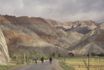 Row of three cyclists on a road leading toward rocky mountain landscape
