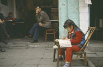 Young girl doing homework at small table outside house watched by adults in open doorway behind with one smoking.