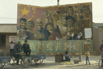 Old Communist poster on hoarding at road junction with woman selling fruit below. Child and two men on stationary bicycle driven carts.