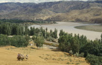 View over river bend in sandy region of north west China.  Group of people on camels in the foreground  mountains behind.