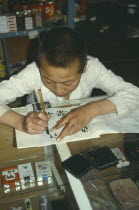 Boy practising writing Chinese characters in shop owned and run by his parents.