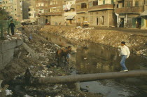 Rubbish in canal with man walking over pipe