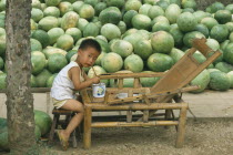 Liitle boy eating from bowl with chopsticks with large pile of watermelons behind.