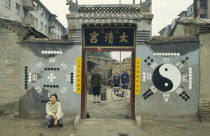Monks or priests seen through the entrance of Daoist temple with trigram and yin yang symbols on the exterior walls and person crouched outside.