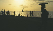 People on the waterfront silhouetted against yellow sunset with dhow at sea in the distance