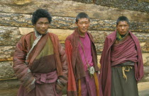 Three Tibetan monks standing against wooden exterior wall of building.