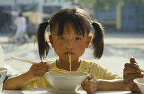 Pony tailed young girl eating noodles from bowl with chopsticks