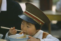 Boy wearing peaked military cap eating noodles from bowl with chopsticks.