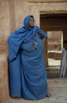 Portrait of woman wearing turquoise robe and head covering standing in front of mud brick building with child framed in doorway behind her. West Africa