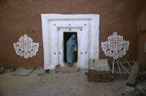 Woman stepping out from doorway with white surround and traditional bas relief motifs on each side of applied gypsum  white and red clay.West Africa