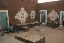 Man preparing food in courtyard of buildings with traditional bas-relief ornamentation of applied gypsum  white and red clay with turquoise painted doors  washing line and goats.West Africa