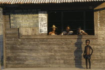 Child standing outside local cinema ticket office with three girls at open window and blackboard on outside wall with film listings in French.