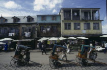 Street scene with washing hanging from buildings above roadside stalls and pousse pousse drivers waiting for customers.  People walking past  some with bags.Tana