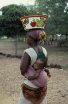 Woman carrying baby in sling on her back and painted bowl on her head in area west of Banfora.