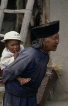 Elderly Tu nationality Lamaist woman of Yellow Hat Buddist sect carrying child on her back.