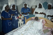 Women surround deceased Ga priestess of the sea god Kanjar laid out on bed during wake.