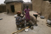 Woman cooking over open stove watched by young girl in small  walled yard with goat kid and thatched mud brick hut behind.West Africa