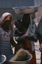 Nigerian woman winnowing grain beside woman carrying child in her arms and large dish on her head.
