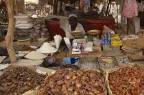 Market trader sitting behind scales and display of dried foodstuffs and other goods.