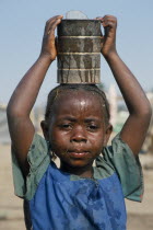 Portrait of Sierra Leonean refugee girl carrying can of water on her head in camp for displaced people.