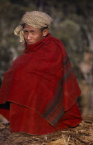 Portrait of a Shan sat down and wrapped in a red blanket with a scarf round his head. Burma