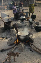 Woman cooking on open fires in village compound with children and goats.
