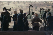 Dogon men carrying carved wooden staffs symbolic of ancient Dogon society performing rooftop dance.