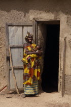 Portrait of Malian mother and breastfeeding baby standing outside open doorway of mud brick building.