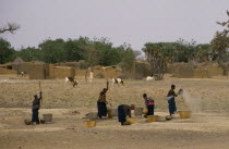 Women pounding millet near Koro with grazing sheep and village buildings beyond.