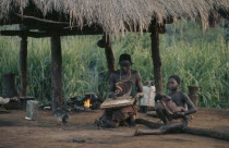 Woman and children sitting beneath thatched shelter in village with pot on open fire behind. Zaire