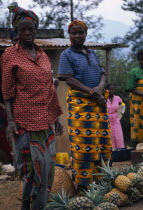 Burundi, Market, Fruit, Women in bright dresses at side of road standing beside their pineapples for sale.