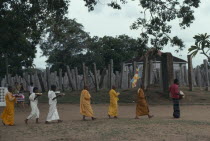 Buddhist monks and nuns carrying puja offerings past columns of the ruins of the Brazen Palace.