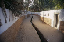 Deep irrigation channel through street between walls partly painted white with triangular cut outs.Ghadamis Gadames