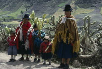 Family on roadside returning home from Palm Sunday Mass holding woven and shaped palm fronds with line of cacti and patchwork fields on hillside behind.