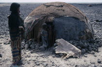 Afar woman carrying child standing outside hut or ari with two young children in the doorway n barren arid landscape.Cushitic speaking pastoral nomads aka Danakil and Adali