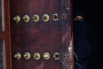 Woman in full chadoor looks out from behind ornate carved and studded door. Unguja