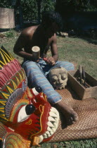 Man carving wooden masks next to a brightly painted example of his work