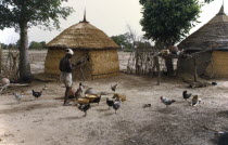 Village scene with thatched mud huts and man feeding chickens.