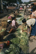 Female vendors and customers at roadside stalls selling herbs and vegetables.