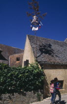 La Roque. Easter decorations attached to pole on exterior of house with two young girls walking past