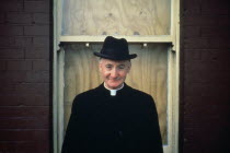 Priest wearing hat in front of boarded up house window.EIREEIREEIREEIREEIRE