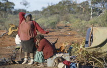 Aboriginal women in an Aborigine camp with one women stretching with her arms over her head next to a woman sat on a chair near a small dog and a tent.