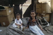 Two Street children sat on sacks smiling surrounded by large cardboard boxes