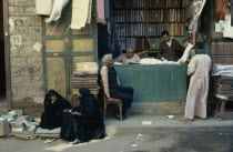 Market scene.  Vendors  customer and women in conversation outside pavement stall selling fabric.