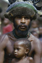 Melpa tribesman wearing hat decorated with fur  feathers and leaves with young child sitting in front of him.