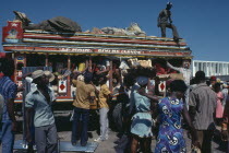 Brightly painted and crowded taptap bus with waiting crowds and food sellers.