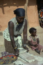 Burji woman grinding grain on large flat stone to produce flour with child sitting beside her.East AfricaBambala