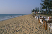 Khao Lak  A couple sat at a table on the sandy beach  people walking along the shore and tall palm trees.