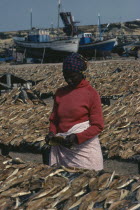 Woman laying out fish to dry with line of moored fishing boats behind.