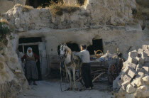 Couple harnessing pony to wooden cart.  Inhabitants of cave dwellings built into rock behind.